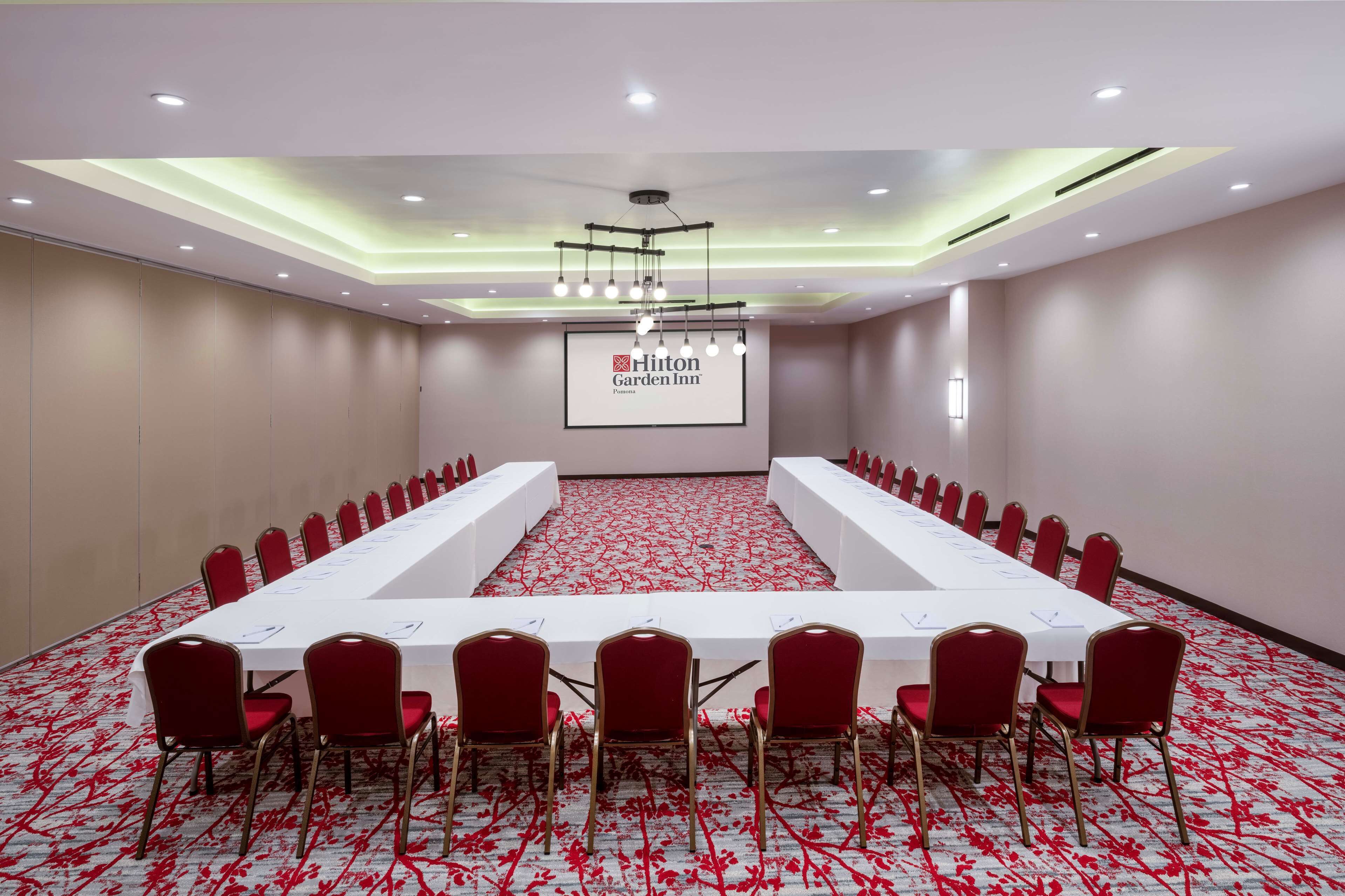A conference room with a long table and red chairs, a projector screen with "Hilton Garden Inn" logo, and a carpet with a red pattern.
