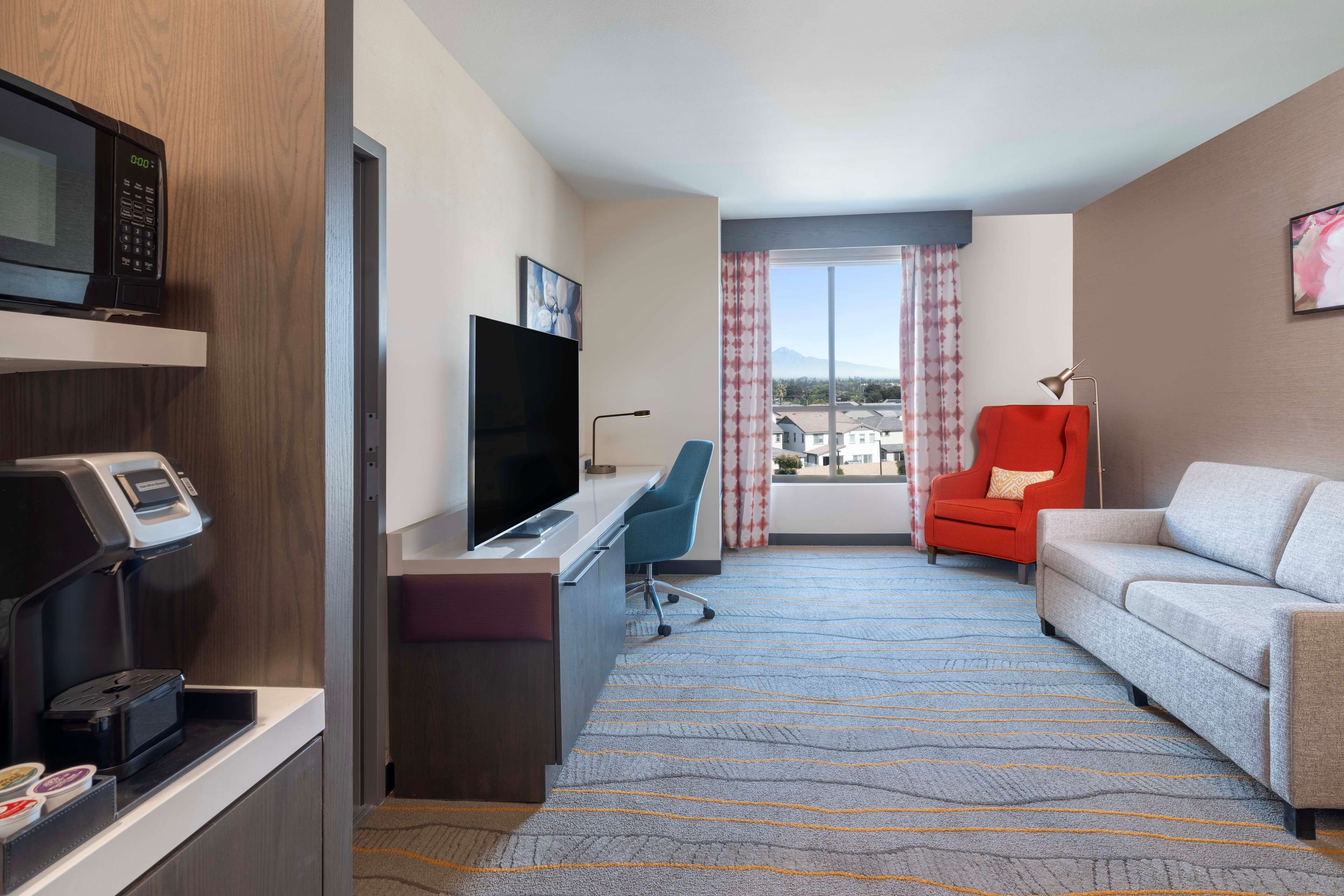 A hotel room with a desk, a television, a microwave, a red armchair, a grey sofa, and a large window with a view of buildings.
