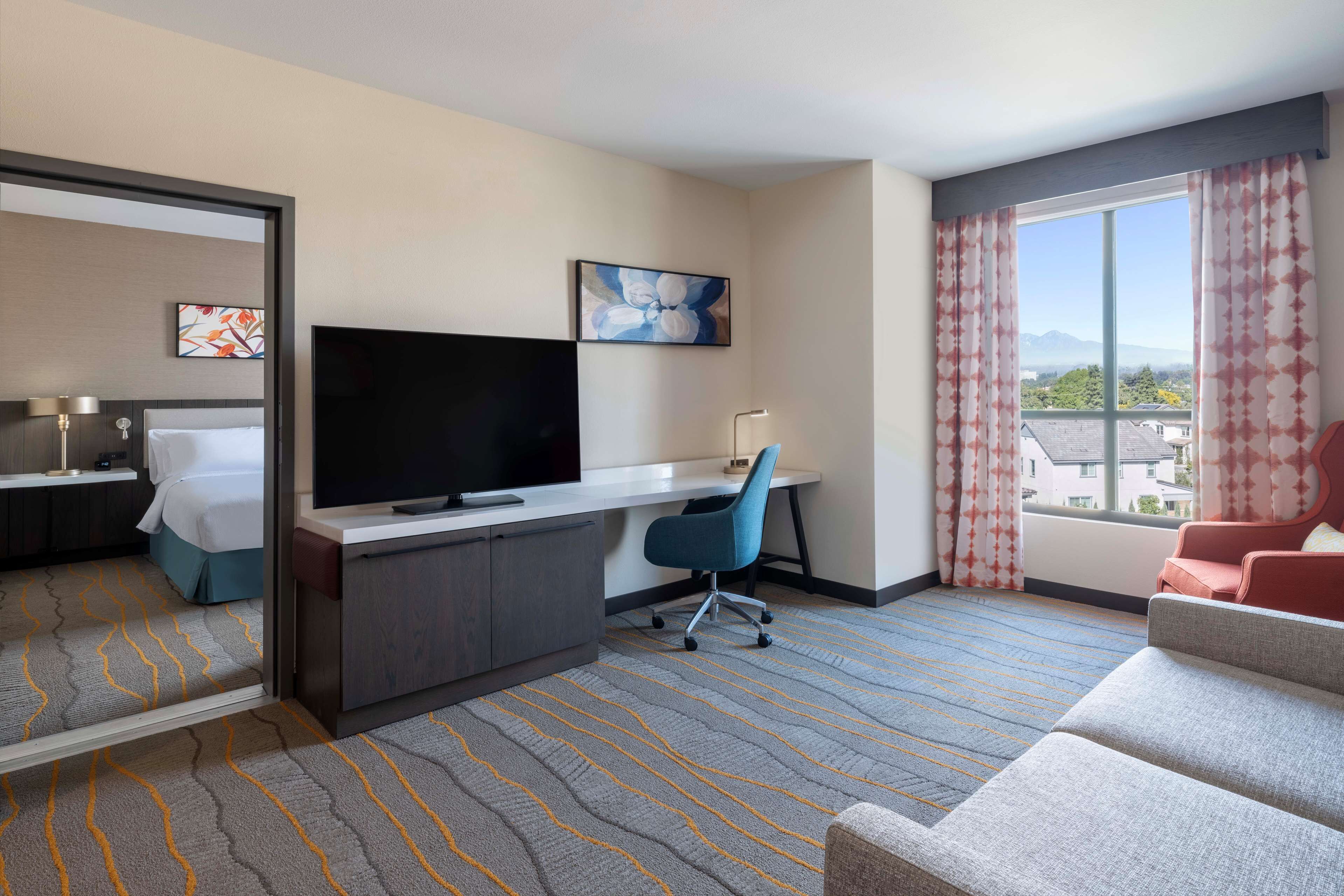 Habitación de hotel con cama, escritorio con televisor, silla de oficina azul, silla roja y ventana grande con vistas.