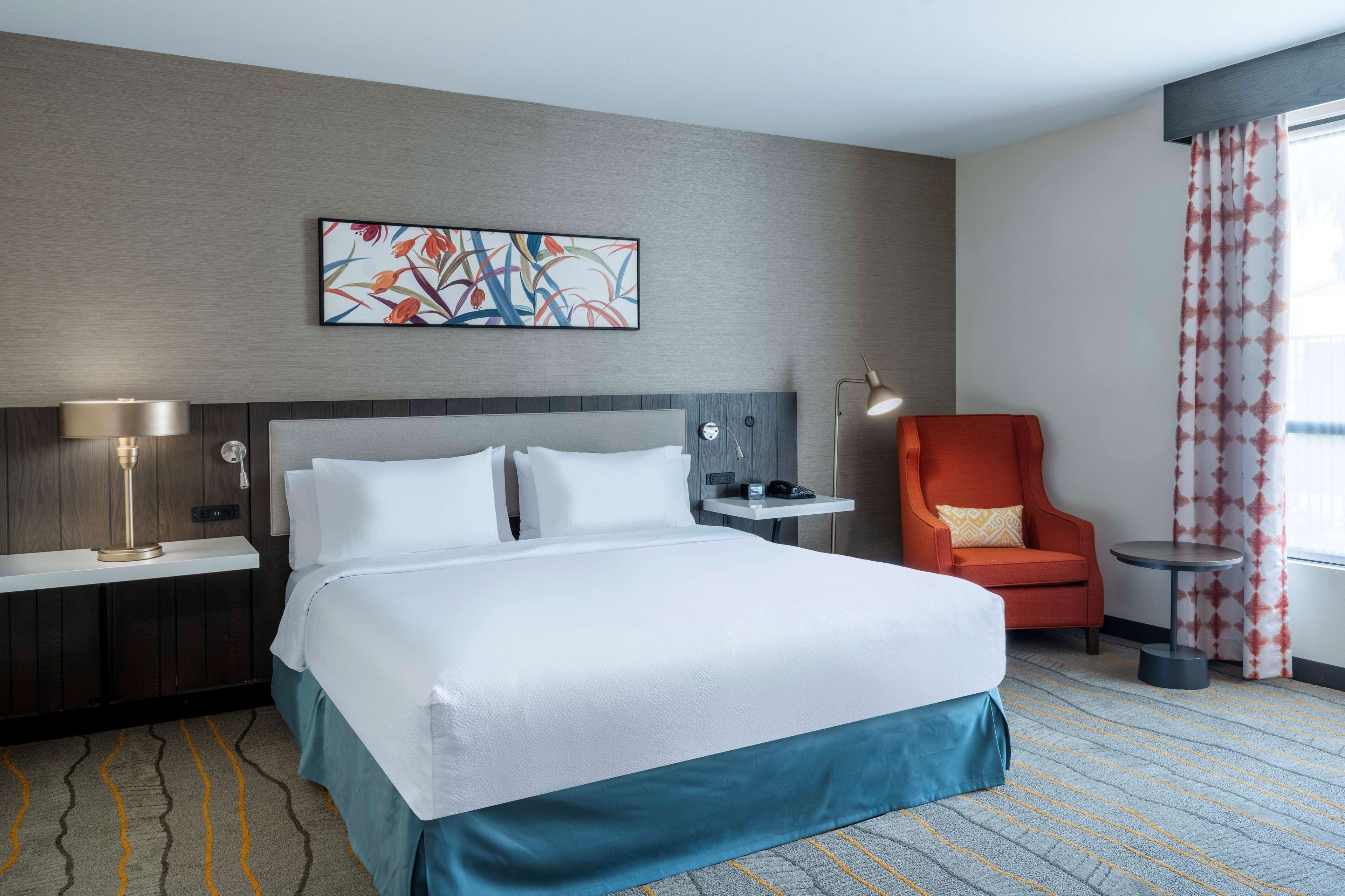A hotel room with a large bed, a desk with a lamp, a chair, and a window with curtains.