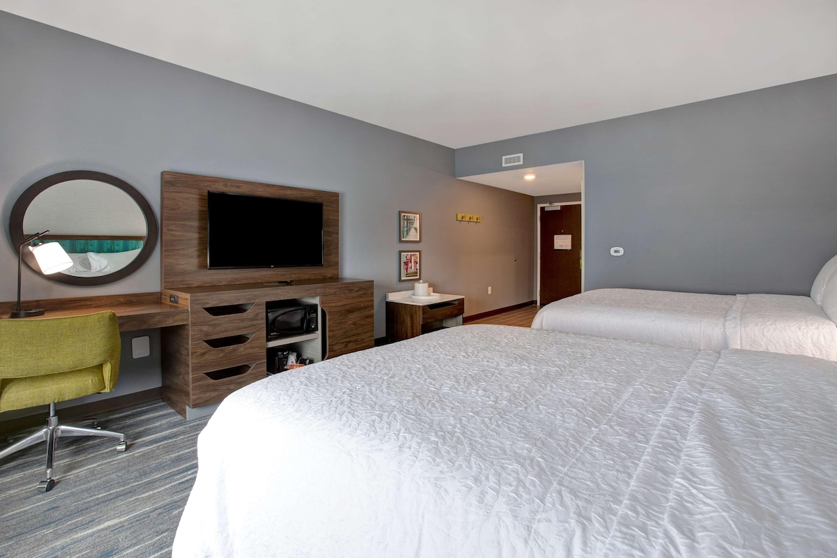 Una habitación de hotel con una cama grande, un escritorio con silla, un televisor y un espejo.