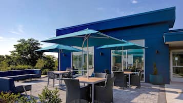 An outdoor patio area with tables and chairs, umbrellas, and a blue building in the background.