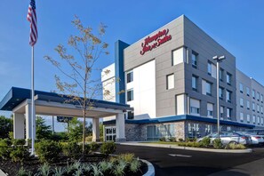 Um hotel Hampton Inn & Suites com design moderno, apresentando entrada acessível, um mastro de bandeira e área ajardinada bem cuidada.