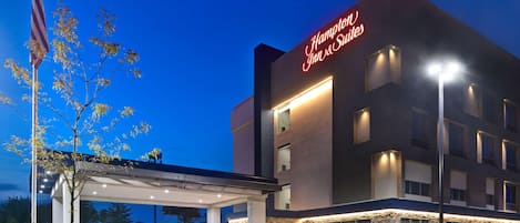 Ein Hampton Inn & Suites Hotel bei blauem Himmel, mit amerikanischer Flagge und beleuchtetem Eingang.