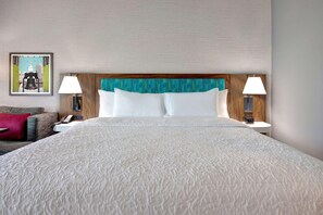 A hotel room with a large bed, two bedside lamps, a nightstand with a phone, a framed picture on the wall, and a red throw pillow on the couch.
