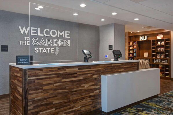 Un comptoir d’accueil avec une pancarte indiquant « BIENVENUE DANS L’ÉTAT DES JARDINS », une autre indiquant « RESPONSABLE DE SERVICE » et une troisième indiquant « HILTON HONORS ».