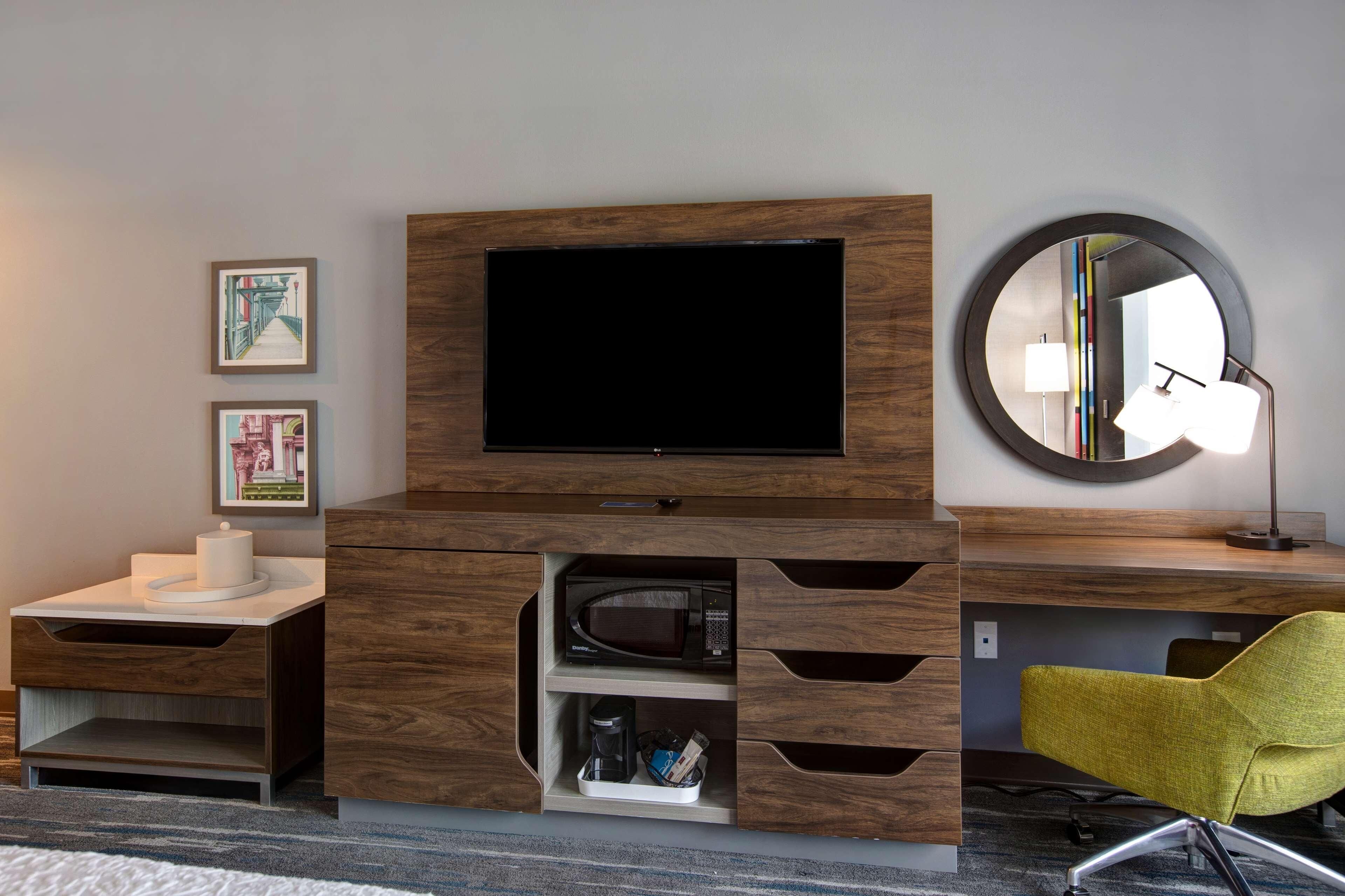 A modern hotel room with a flat-screen TV, a round mirror, a coffee maker, and a bright yellow chair.