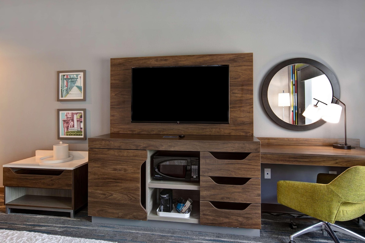 Una habitación de hotel moderna con un escritorio de madera, un televisor de pantalla plana, un espejo redondo y una silla amarilla.