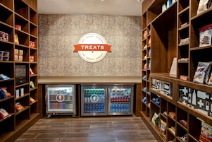 Sección de snacks y artículos de conveniencia con un cartel que dice 'Treats', dispensadores refrigerados para bebidas y estantes llenos de diversos snacks y papas fritas.