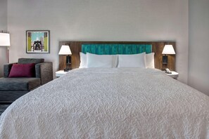 A hotel room with a large bed, two bedside tables with lamps, a grey sofa with a red pillow, and a framed picture on the wall.