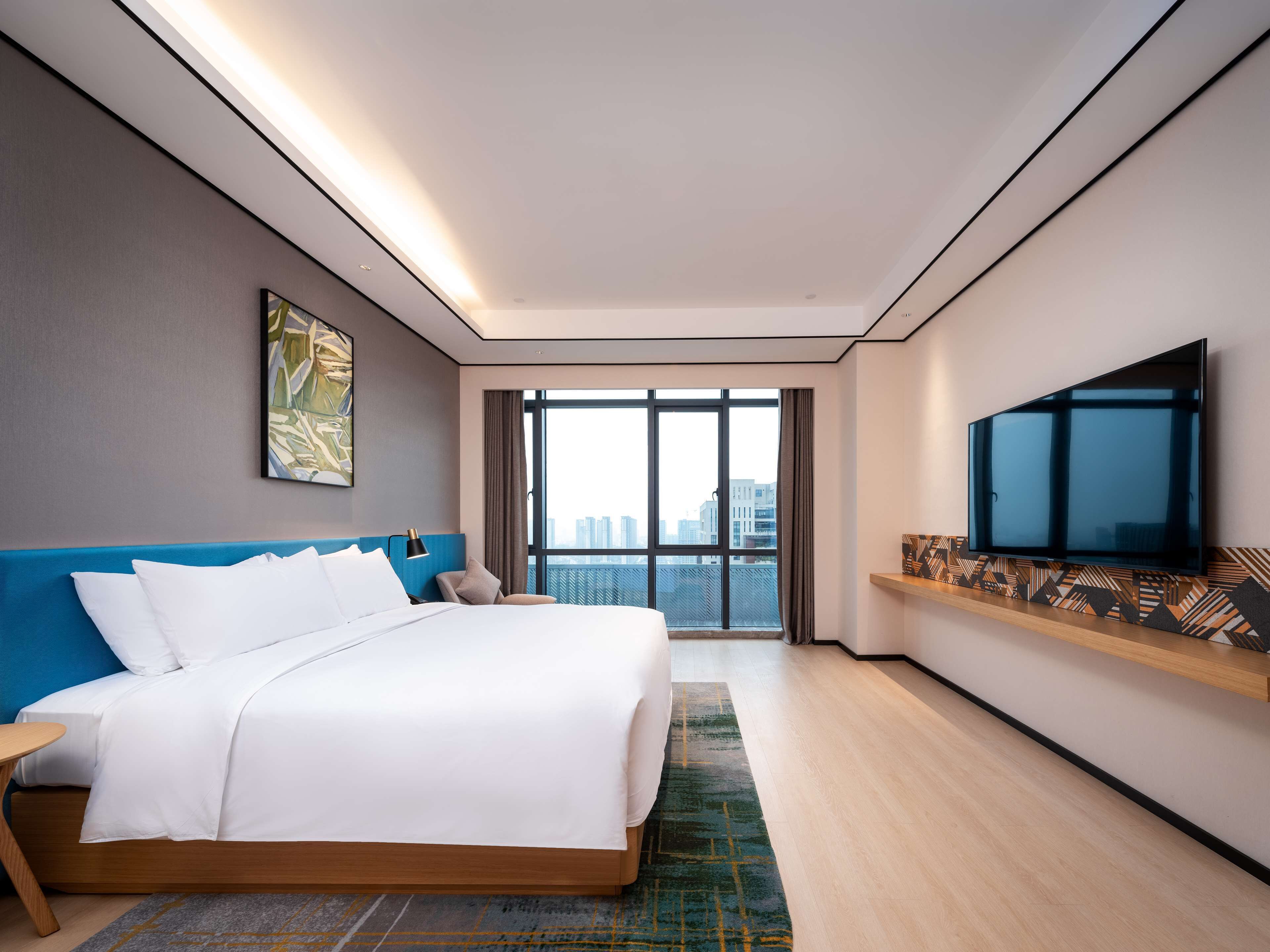 A modern hotel room with a large bed, a flat-screen TV, a city view, and a minimalist design.