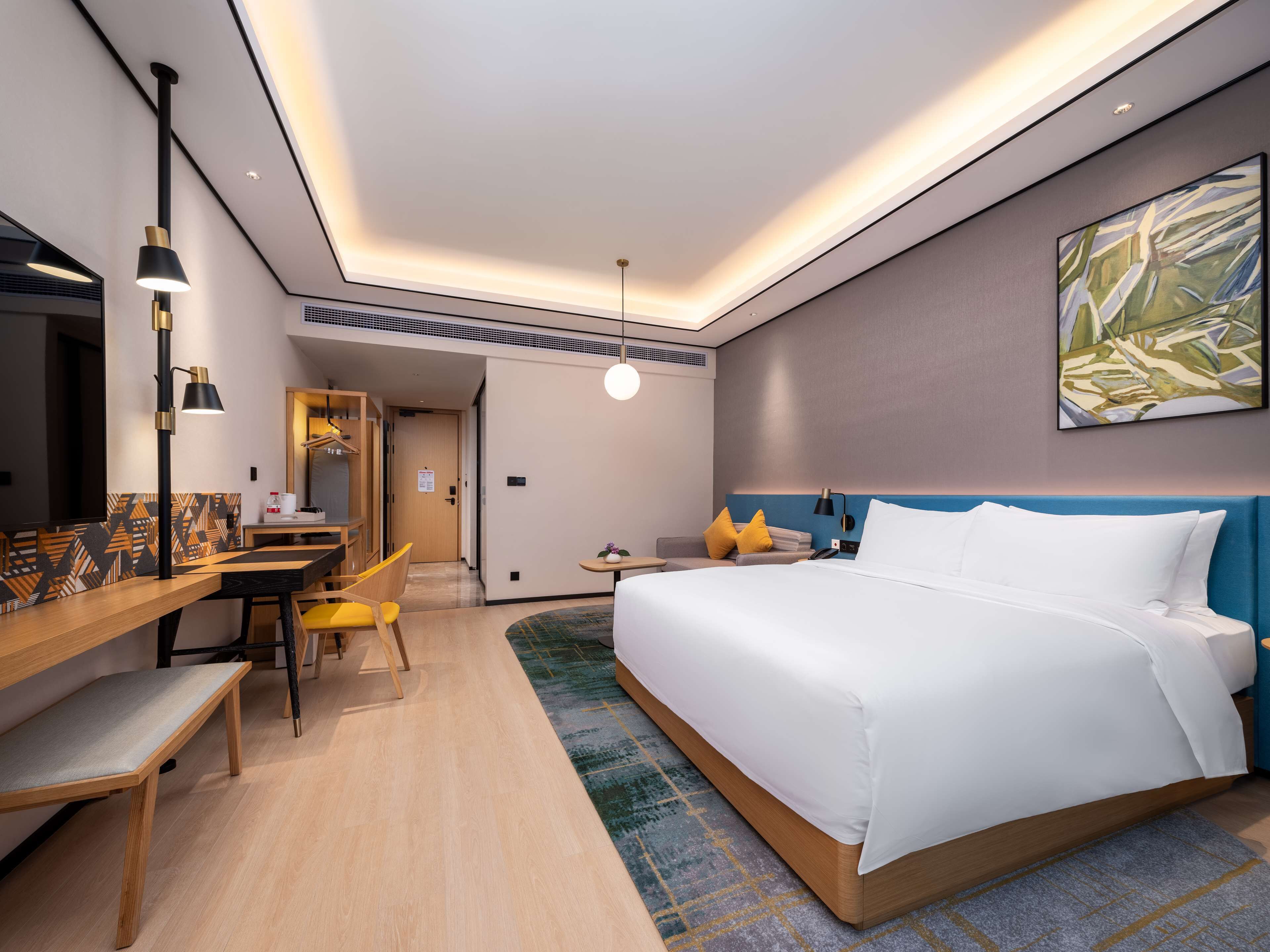A modern hotel room with a large bed, a desk, and a chair. There is a painting on the wall and a ceiling light.