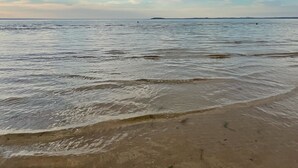 Una platja amb aigua tranquil·la, sorra daurada i un cel nuvolós.