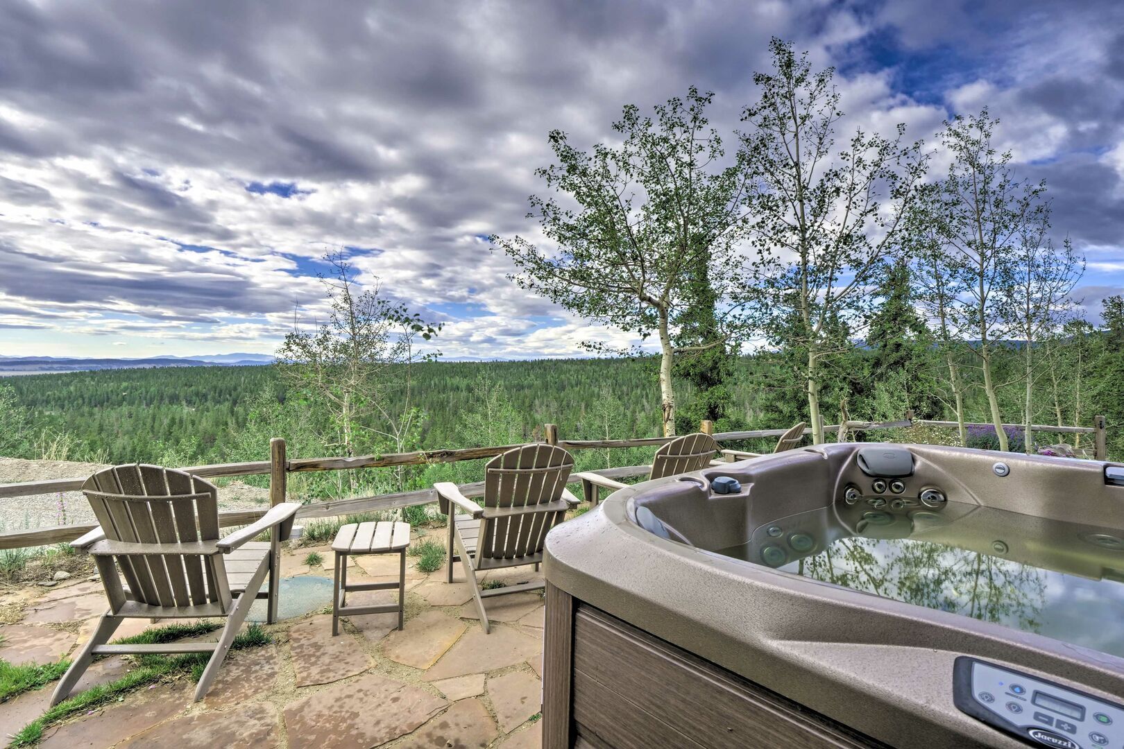 Un bain à remous doté d’un panneau de commande, entouré de chaises en bois et d’une table, et offrant une vue sur un paysage boisé.