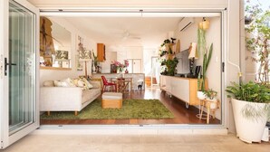 An open-plan living area with a sofa, dining table, and chairs, a kitchen area with a fridge and plants, and a large window.