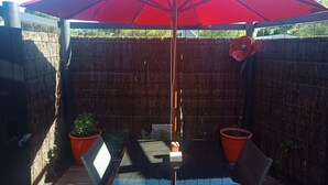 Une terrasse avec un parasol rouge, deux plantes en pot et une table en verre.