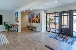 A modern interior with a wooden floor, a green chair, a small table, and a bench. There are three framed pictures on the wall and a potted plant near the entrance.