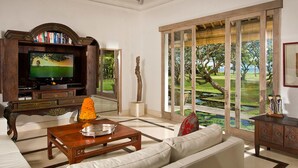 A living room with a television, a coffee table, a sofa, a wooden cabinet, and a view of a garden through a glass door. - 5-Star Beachfront Villa, Bali Villa 1014 (Bali)