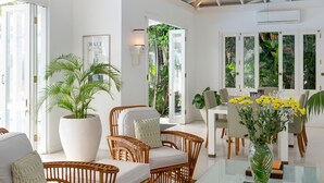 A bright, airy room with wicker furniture, a large potted plant, and a dining area with a view of lush greenery.