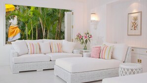 A modern living room with a white sectional sofa, a white ottoman, and a wicker coffee table. There is a painting of a tropical scene on the wall, and a vase with pink flowers on a side table.