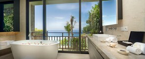 A modern bathroom with a large bathtub, a sink, and a mirror. There is a balcony with a view of the ocean and palm trees. - Bali Villa 1082: Cliffside Luxury with Ocean Views (Bali)