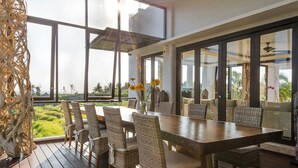 Un comedor con una larga mesa de madera y sillas de mimbre, ventanas amplias que ofrecen vistas a zonas verdes y edificios, y una estructura tejida decorativa a la izquierda.