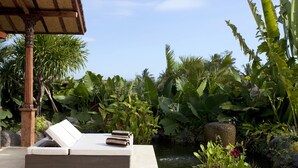 A serene outdoor lounge area with a wicker sofa and a small pond, surrounded by lush tropical plants and a wooden pergola.