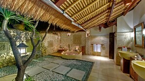 Een wellnessruimte met een stenen muur, rieten dak, stenen lantaarn, een rond bubbelbad en een grote wastafel.