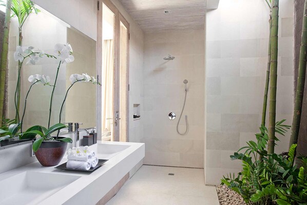 A modern bathroom with a large mirror, white sink, and shower. There are orchids and other plants around.
