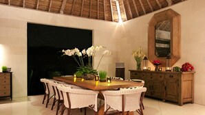 A dining area with a large wooden table surrounded by wicker chairs, a wooden cabinet with decorative items, and a ceiling with a woven design. - Serenity by the Shoreline – Bali Villa 1048 (Bali)