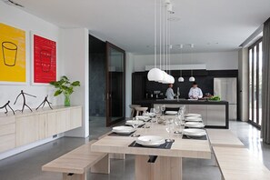 A modern dining area with a long table set for a meal, a kitchen area with two chefs, and a large window providing natural light.