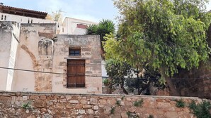 Edificio de piedra con puerta de madera y una ventana pequeña, rodeado de un muro de piedra y vegetación.