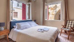 Una cama bien hecha con sábanas blancas, una silla con cojín escocés y una ventana con vistas a un muro de piedra.
