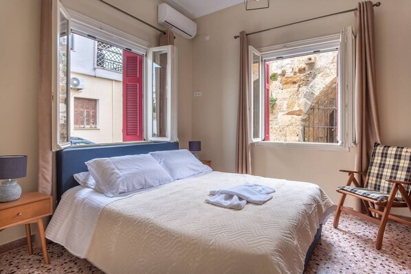 Una cama bien hecha con sábanas blancas, una silla con cojín escocés y una ventana con vistas a un muro de piedra.