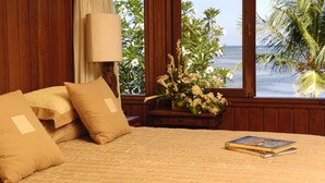 A bedroom with a bed, pillows, a bedside table with a lamp, a vase of flowers, and a view of the ocean through large windows.