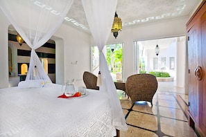 A room with a bed under a canopy, a table with a pitcher and glasses, wicker chairs, and a view of the outdoors.