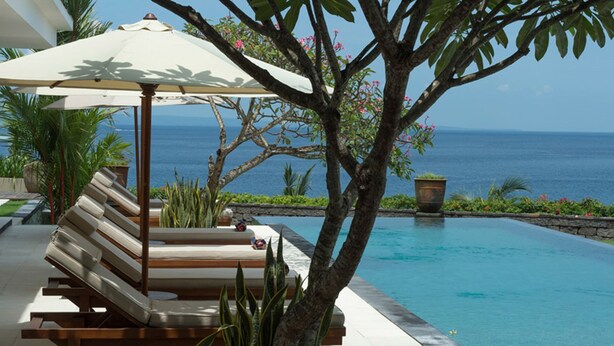 A poolside area with a lounge chair and umbrella, surrounded by tropical plants and a view of the ocean.