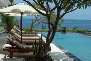 A poolside area with a lounge chair and umbrella, surrounded by tropical plants and a view of the ocean.