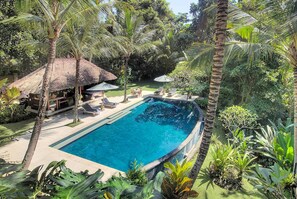 A swimming pool surrounded by palm trees and tropical plants, with a thatched-roof structure and lounge chairs. - Luxury Hillside Retreat with Private Pool – Bali Villa 1049 (Bali)