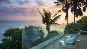 A glass-enclosed balcony with two white lounge chairs, overlooking a body of water and palm trees, with a clear sky above. - Cliffside Retreat with Pool & Ocean Views – Bali Villa 1065 (Bali)