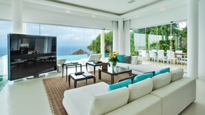 A modern living room with a large flat-screen TV, a white sofa with teal cushions, a coffee table, and a dining area with white chairs and a glass table. - Cliffside Retreat with Pool & Ocean Views – Bali Villa 1065 (Bali)