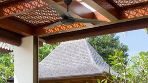 Uma área de estar externa com móveis de vime e almofadas, um gazebo com telhado de palha e uma piscina ao fundo.