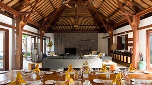 Uma sala de jantar espaçosa, com teto alto e vigas de madeira, um amplo sofá ao fundo e uma mesa de jantar posta para refeição.