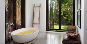 Ein Badezimmer mit einer großen weißen Badewanne, gefüllt mit gelben Badesalzen, einer Holzleiter und Blick auf einen üppigen, grünen Wald durch ein großes Fenster.