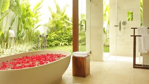Une salle de bain dotée d’une grande baignoire remplie de pétales de roses rouges, d’un tabouret en bois, d’une cabine de douche en verre et offrant une vue sur une végétation luxuriante à l’extérieur.