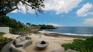 Un espace de détente aménagé sur la plage, avec un canapé incurvé et un foyer en pierre, offrant une vue sur l’océan et le rivage rocheux.
