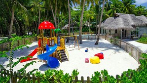 Une aire de jeux au bord de la plage, avec un toboggan, des balançoires et un abri à toit de chaume.