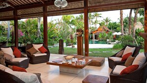 A spacious indoor area with wicker furniture, a coffee table, and a view of a pool and tropical garden. - Secluded Comfort, Steps from the Beach – Bali Villa 1096 (Bali)
