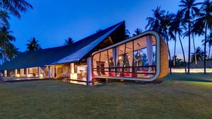 A modern, curved building with large glass windows and a sloped roof, surrounded by palm trees. - Ocean View Luxury Escape Lombok Villa 1133 (Lombok)