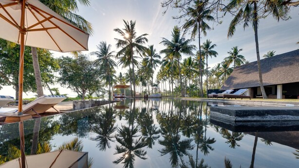 A serene pool area with lounge chairs and umbrellas, surrounded by tall palm trees and a modern building. - Ocean View Luxury Escape Lombok Villa 1133 (Lombok)